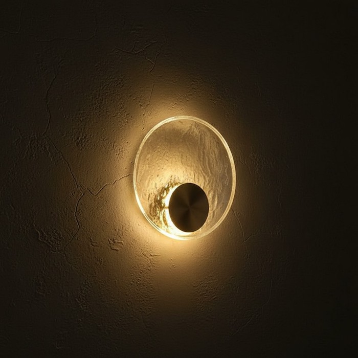 Side view of a gold circular sconce showing physical depth and light halo on a dark wall.